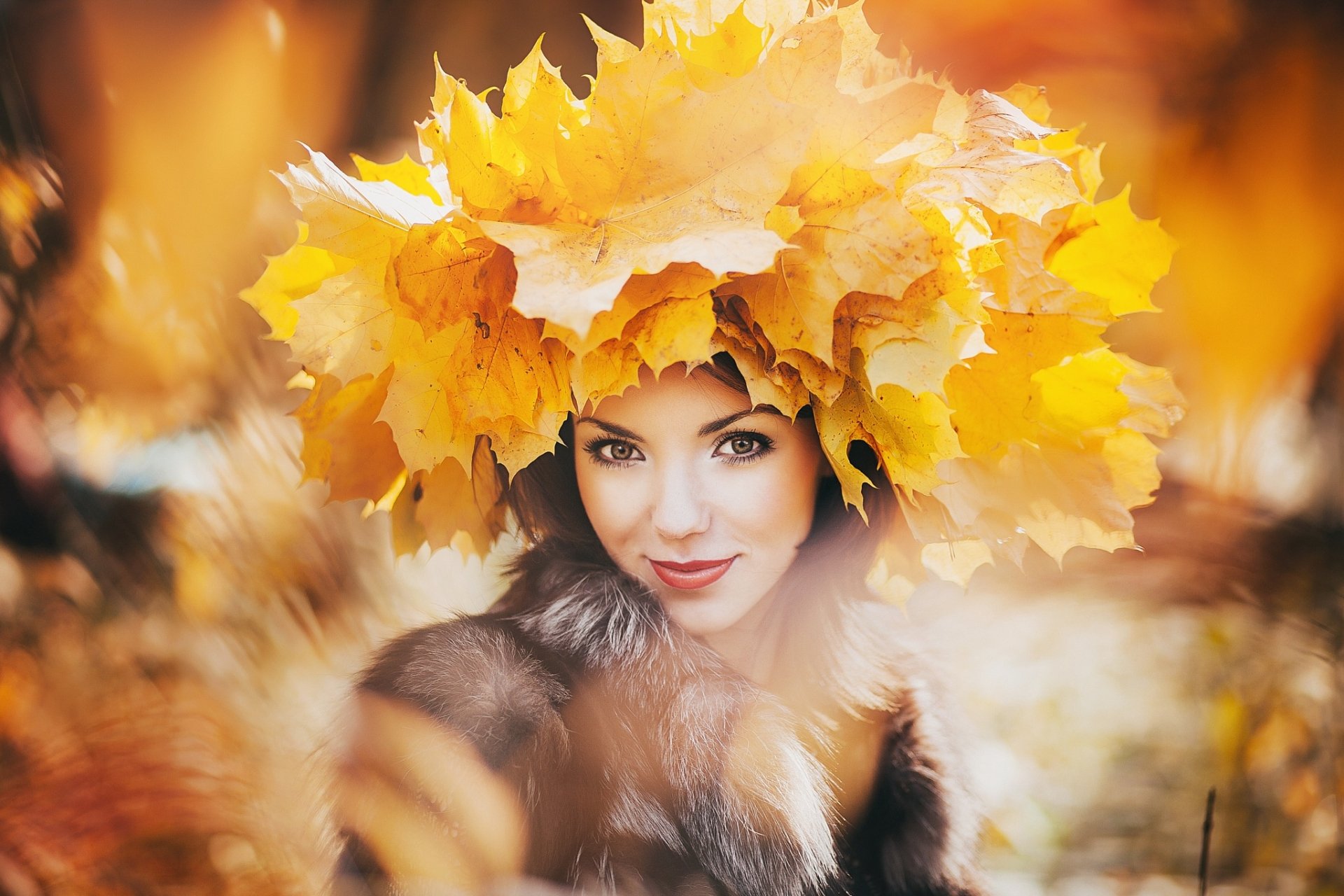 A smiling woman with hazel eyes and lipstick wears a crown of yellow leaves, surrounded by soft fur and blurred autumnal background in this HD desktop wallpaper.
