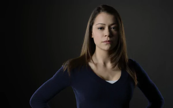 HD desktop wallpaper of a brunette woman with brown eyes standing against a dark backdrop, hands on hips and a calm, confident expression.
