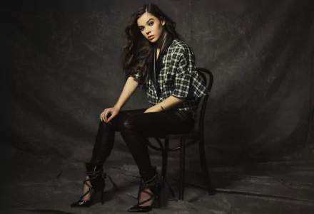 Hailee Steinfeld, a brunette American actress, poses confidently on a chair, wearing a stylish plaid jacket, against a dark backdrop in this HD desktop wallpaper.