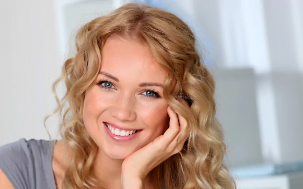 A smiling blonde woman with blue eyes and curly hair rests her face on her hand in this HD desktop wallpaper.