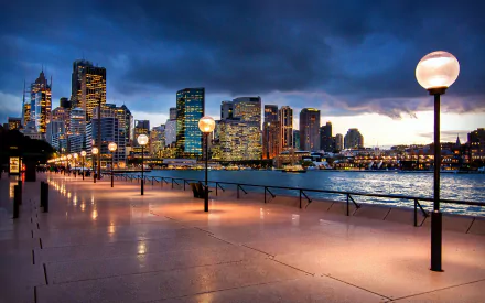 A serene view of Sydney Harbour at Circular Quay, showcasing modern buildings and glowing lamp posts along the waterfront promenade under a dramatic sky.