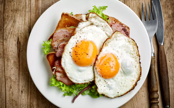 A 4K Ultra HD desktop wallpaper showing a meal with two sunny-side-up eggs served on toast with ham and lettuce on a white plate, placed next to a fork and knife.