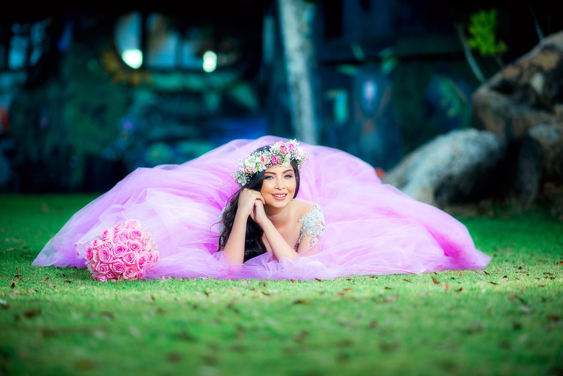 A smiling brunette model lies on grass in a pink dress, wearing a floral wreath and holding a bouquet, captured in this vibrant HD desktop wallpaper.