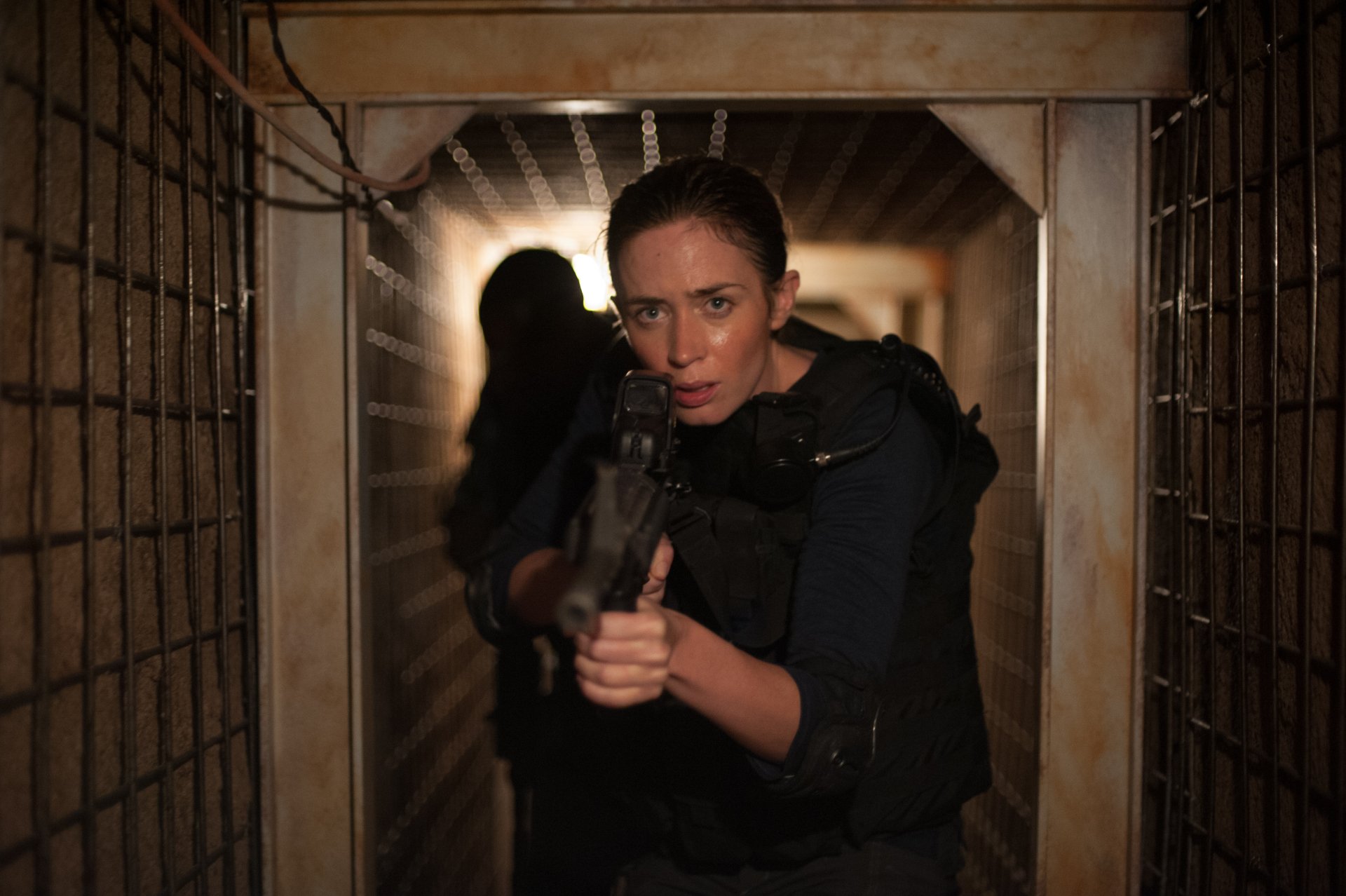 4K Ultra HD movie still (Sicario): a woman in tactical gear aims a rifle down a narrow corridor, intense cinematic lighting for a PC desktop wallpaper.