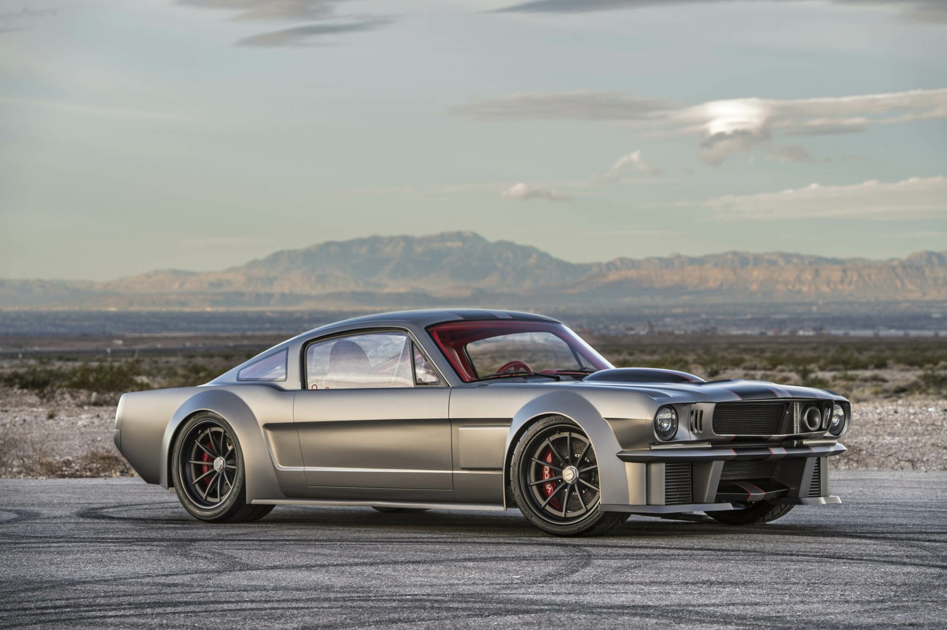 HD wallpaper of a sleek silver Ford Mustang hot rod parked on a scenic desert road, with mountains and a partly cloudy sky in the background.