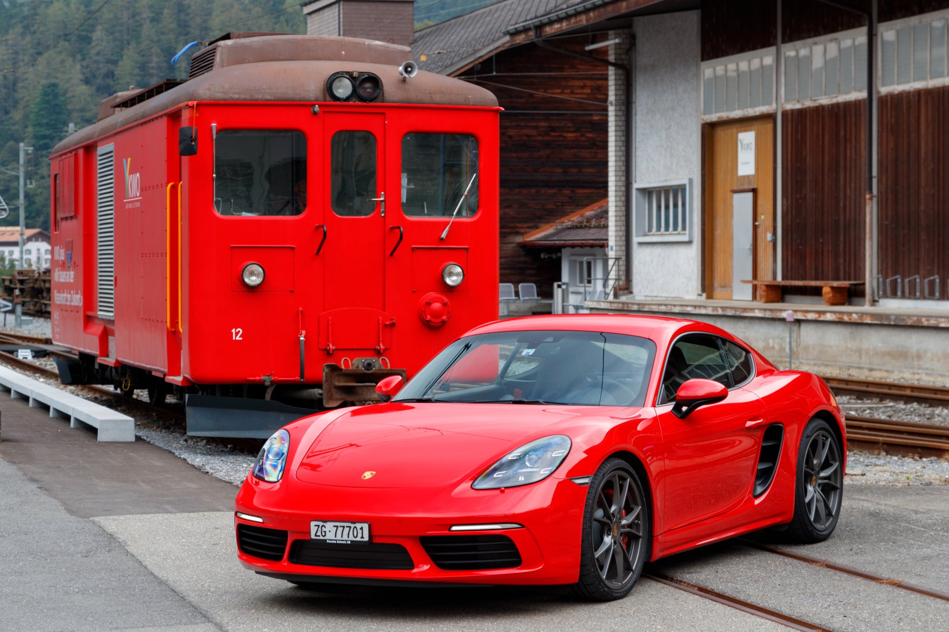 Red Porsche Cayman S sports car parked beside a red train at a station — 4K Ultra HD PC desktop wallpaper background featuring train, car and vehicle.
