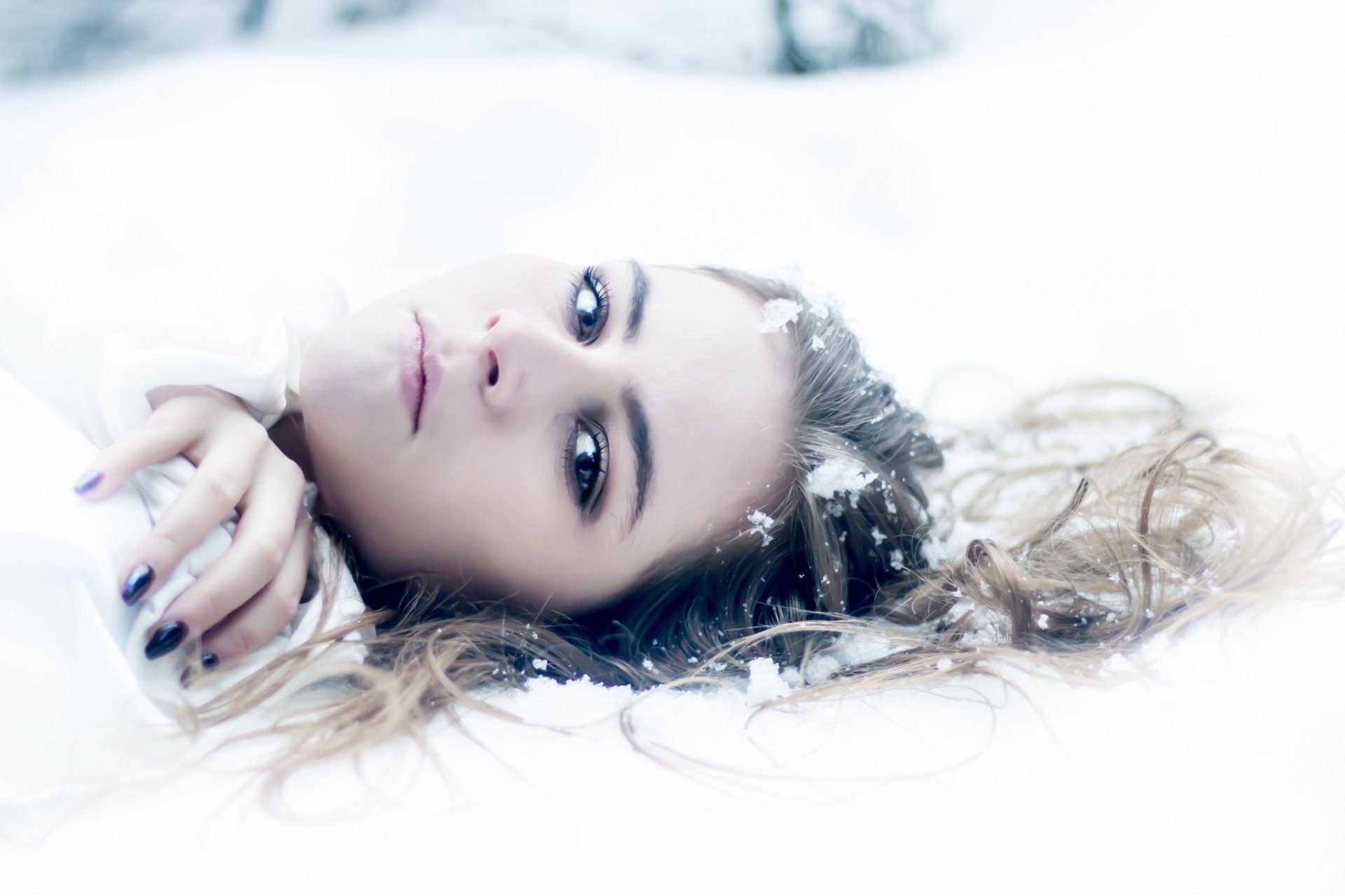 A brunette woman with hazel eyes lies in the snow, her face serene and framed by scattered hair, captured in high-definition detail for a striking desktop wallpaper.