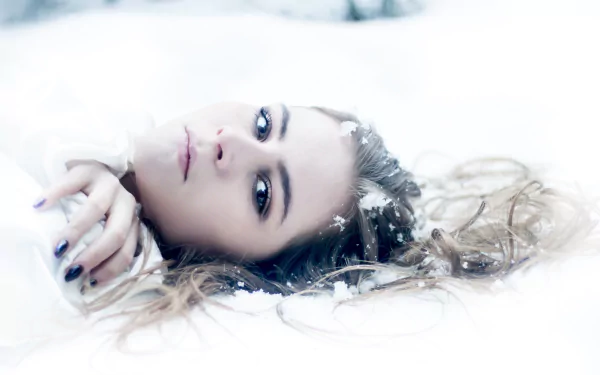 A brunette woman with hazel eyes lies in the snow, her face serene and framed by scattered hair, captured in high-definition detail for a striking desktop wallpaper.