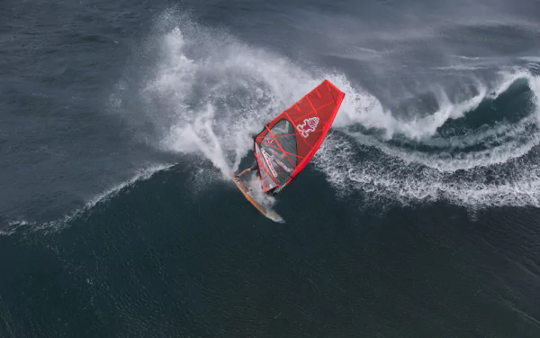 A windsurfer riding a large ocean wave, captured in dynamic action with vibrant red sail, presented as a 4K Ultra HD sports wallpaper and background.
