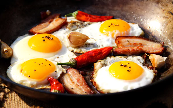Four sunny-side-up eggs fried with slices of bacon, whole red peppers, and garlic cloves in a pan, captured in 4K Ultra HD for a PC desktop wallpaper.