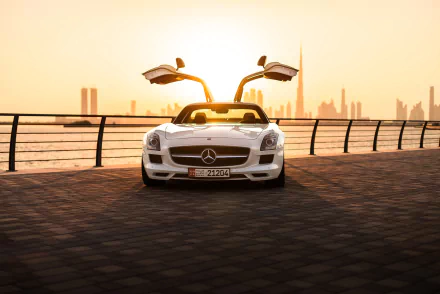White Mercedes‑Benz SLS AMG with gullwing doors open on a waterfront promenade at sunset, city skyline behind — HD PC desktop wallpaper/background