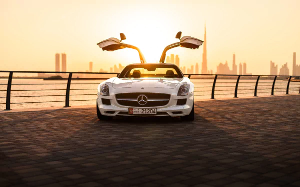 White Mercedes‑Benz SLS AMG with gullwing doors open on a waterfront promenade at sunset, city skyline behind — HD PC desktop wallpaper/background