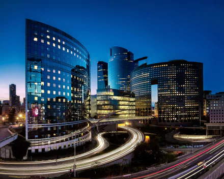 Nighttime time-lapse of La Défense in Paris, showcasing illuminated buildings and winding roads with light trails in this HD cityscape wallpaper.