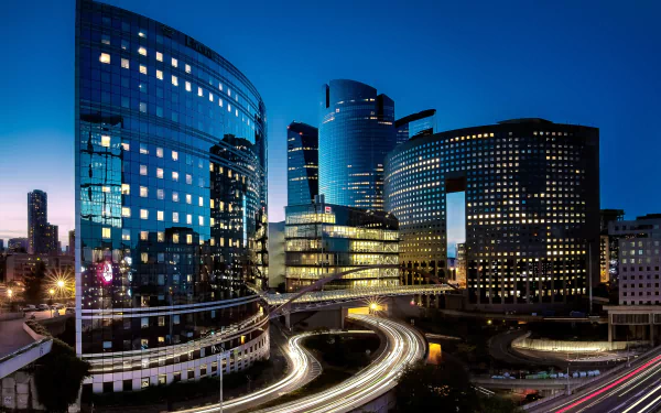 Nighttime time-lapse of La Défense in Paris, showcasing illuminated buildings and winding roads with light trails in this HD cityscape wallpaper.