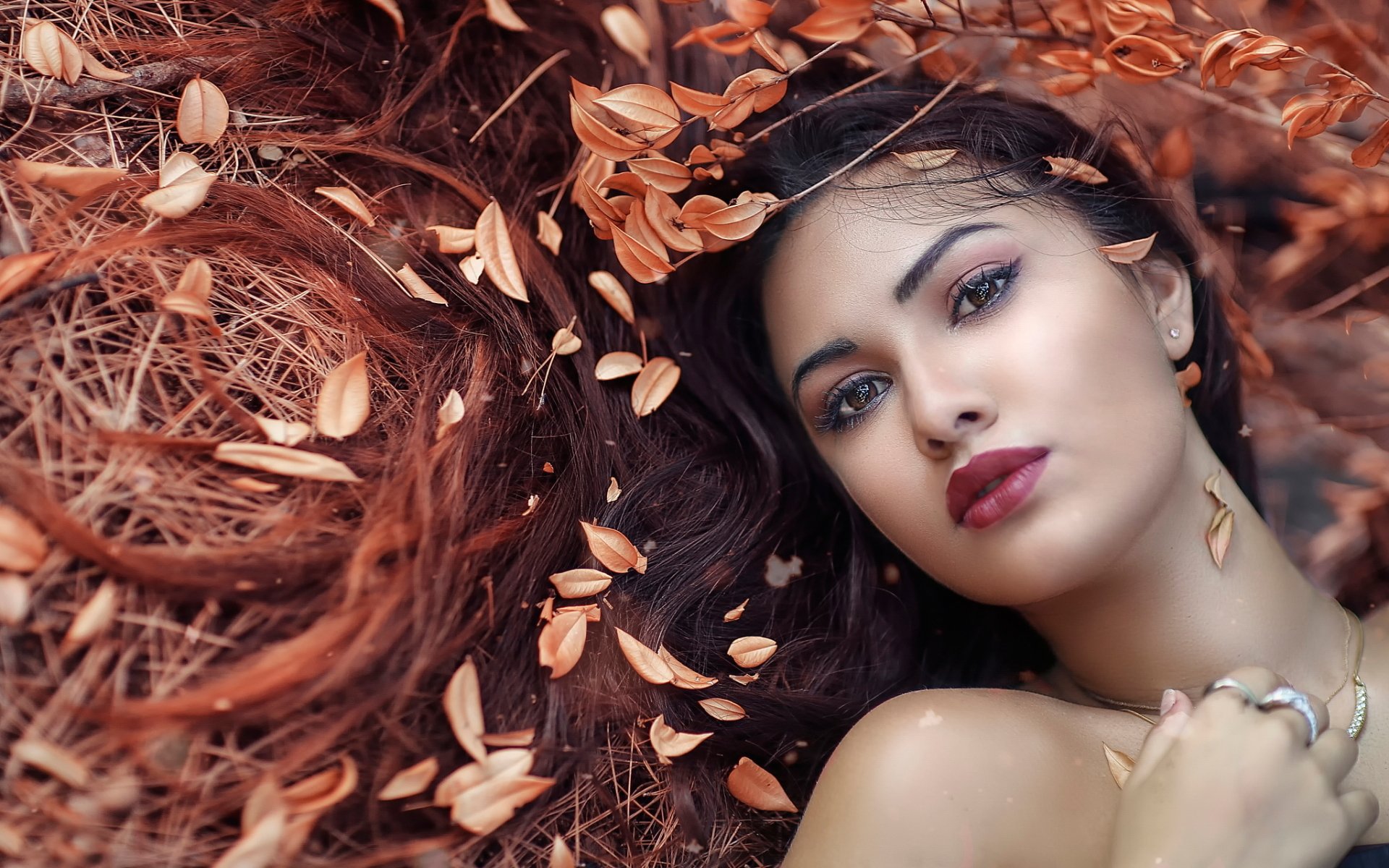A brunette model with brown eyes and lipstick lies down among autumn leaves, creating a captivating fall-themed HD desktop wallpaper and background.