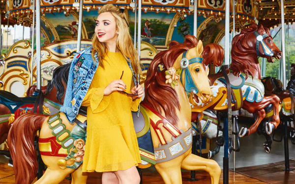 American actress Peyton List with brown eyes and blonde hair smiles in a yellow dress and denim jacket, standing by colorful carousel horses in an HD desktop wallpaper.