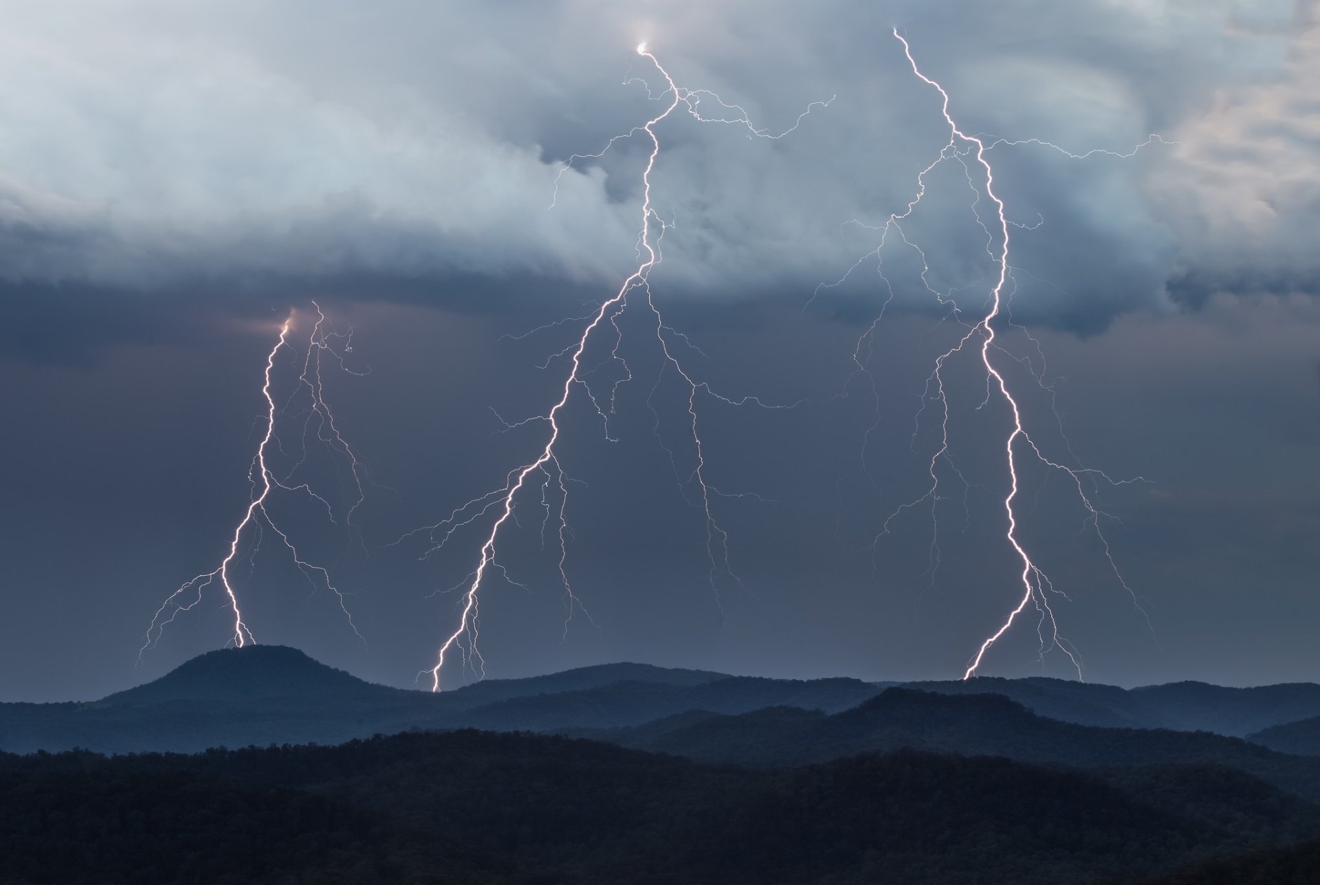 HD desktop wallpaper capturing multiple lightning strikes over a mountainous landscape beneath dramatic cloudy skies, showcasing nature's powerful beauty.