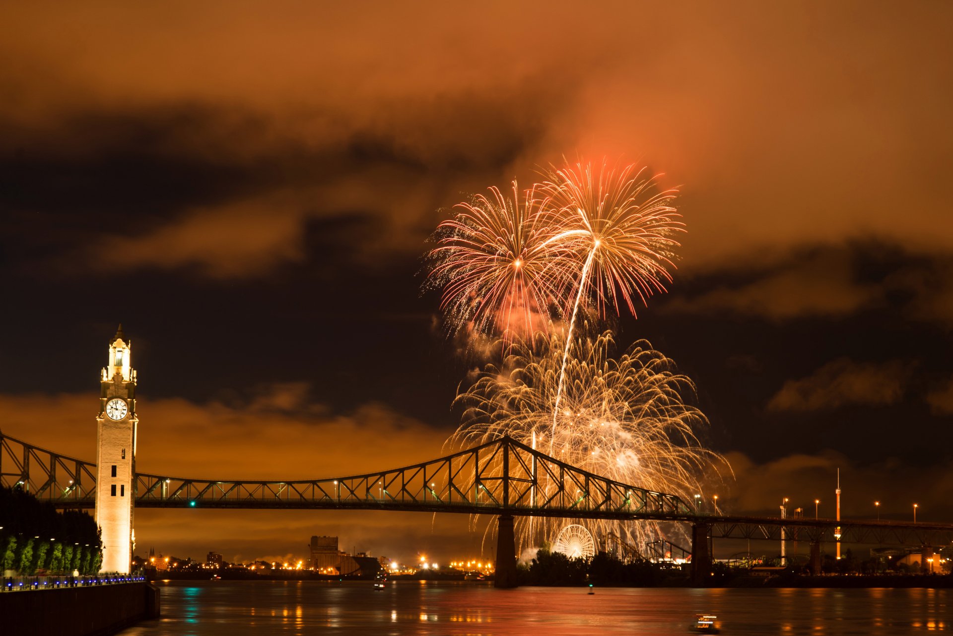 4K Ultra HD wallpaper of Montreal's illuminated bridge over a river at night with vibrant orange fireworks lighting up the sky in Canada.