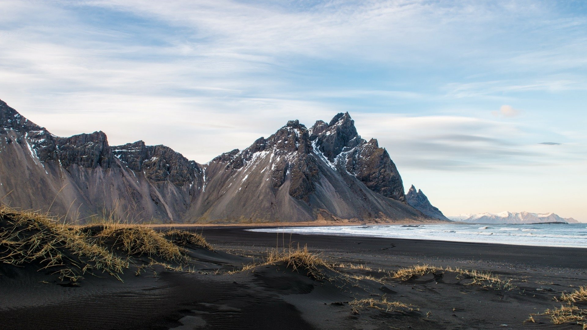 Vestrahorn Mountain in Iceland rises sharply against a cloudy sky, with black volcanic sand and sparse grass in the foreground, showcasing striking natural beauty.