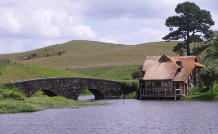 The Lord of the Rings Hobbiton scene: thatched house by a stone bridge over a river with rolling Comarca hills — HD PC desktop wallpaper.