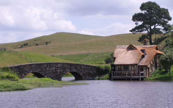 The Lord of the Rings Hobbiton scene: thatched house by a stone bridge over a river with rolling Comarca hills — HD PC desktop wallpaper.
