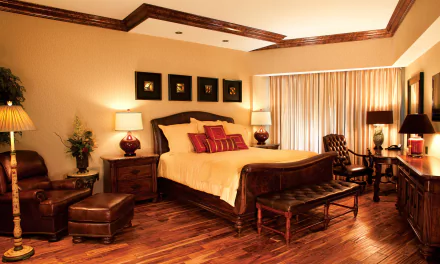 HD desktop wallpaper of a cozy bedroom featuring a bed with cushions, wooden chair, ottoman, and warm lighting in a well-decorated man-made room.