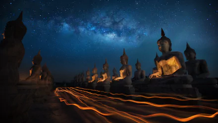 Nighttime HD wallpaper of illuminated Buddha statues in Thailand beneath a starry sky with a vibrant Milky Way and dynamic light trails from a time-lapse capture.