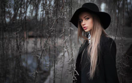 A blonde woman with striking blue eyes wears a black hat and coat, standing amid branches with a blurred depth of field in this HD desktop wallpaper.