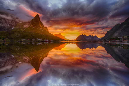 Stunning HD desktop wallpaper of a sunset over a lake in Lofoten, Norway, featuring a mountain, village, and vibrant sky perfectly reflected in the calm water, creating a serene and picturesque landscape.