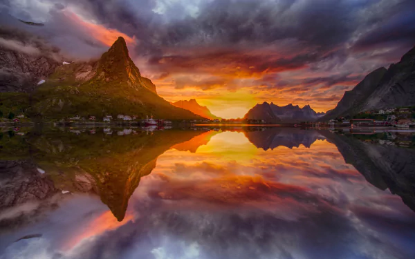 Stunning HD desktop wallpaper of a sunset over a lake in Lofoten, Norway, featuring a mountain, village, and vibrant sky perfectly reflected in the calm water, creating a serene and picturesque landscape.