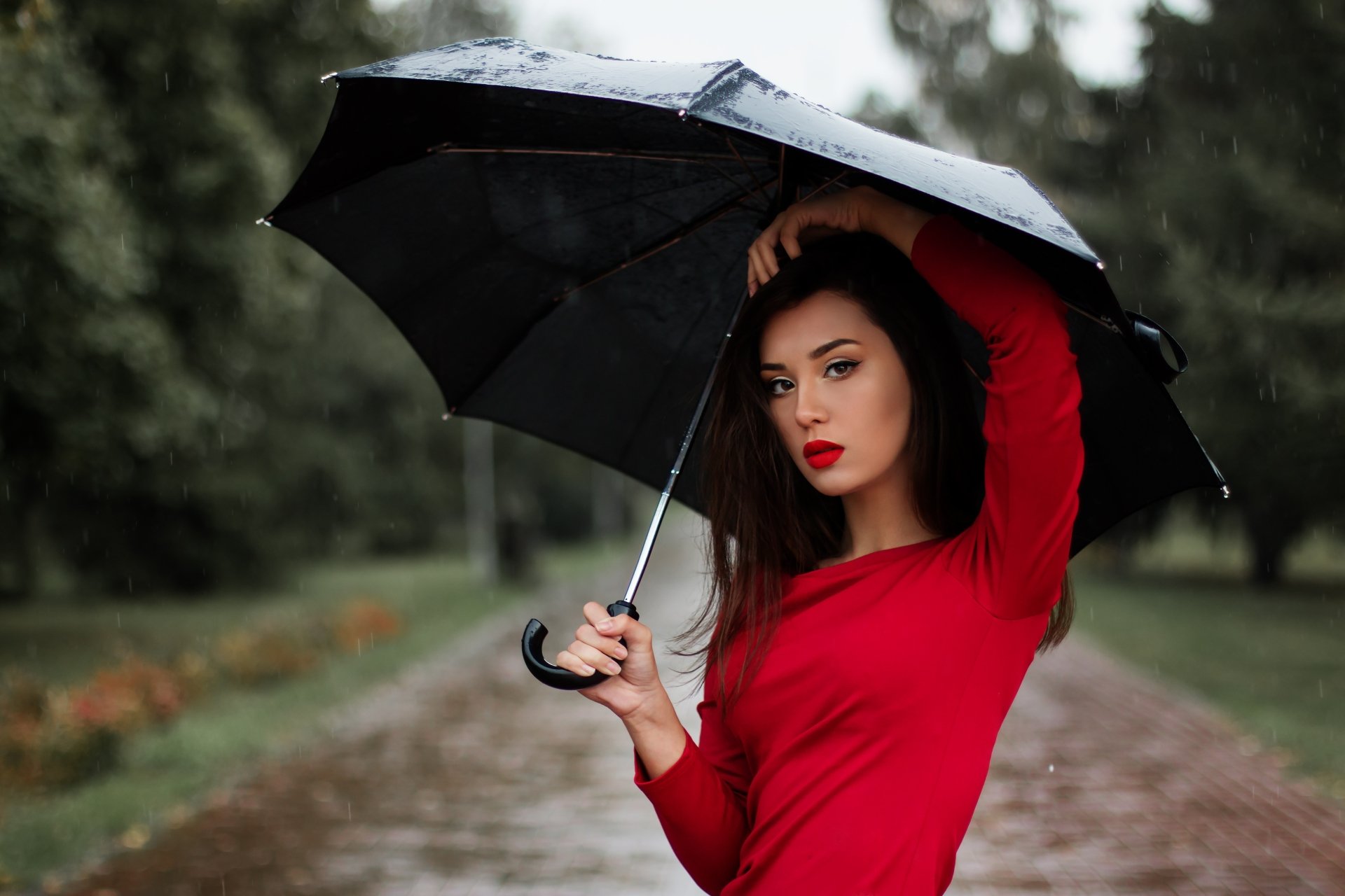 Brunette model with brown eyes and red lipstick holds a black umbrella in the rain, wearing a red top — 5K Ultra HD PC desktop wallpaper background.