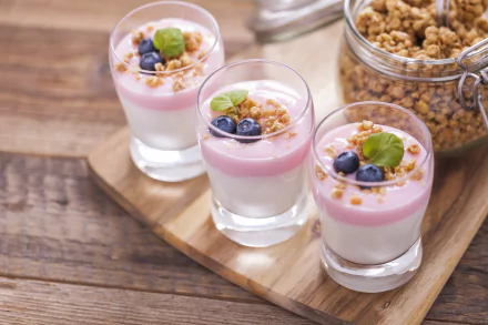 Close-up of three glasses with layered blueberry yogurt dessert topped with granola and fresh blueberries, set next to a jar of muesli on a wooden board.