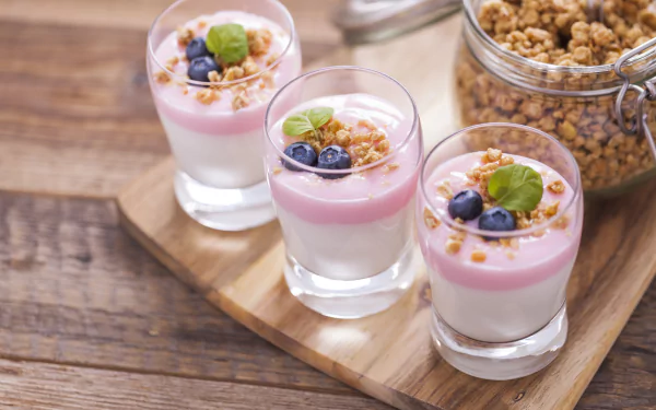 Close-up of three glasses with layered blueberry yogurt dessert topped with granola and fresh blueberries, set next to a jar of muesli on a wooden board.