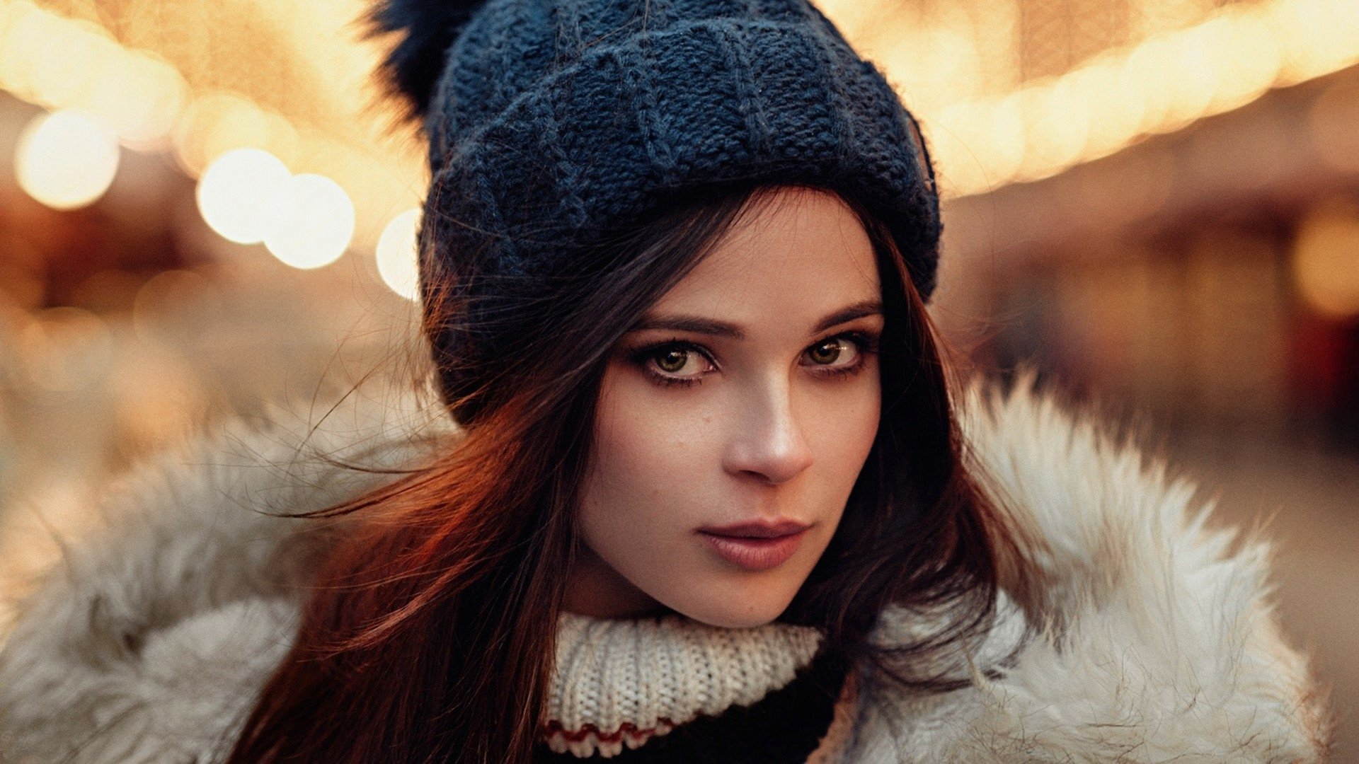 A striking model with red hair and hazel eyes wears a cozy hat, set against a bokeh background. This HD image captures her captivating expression and stylish look.