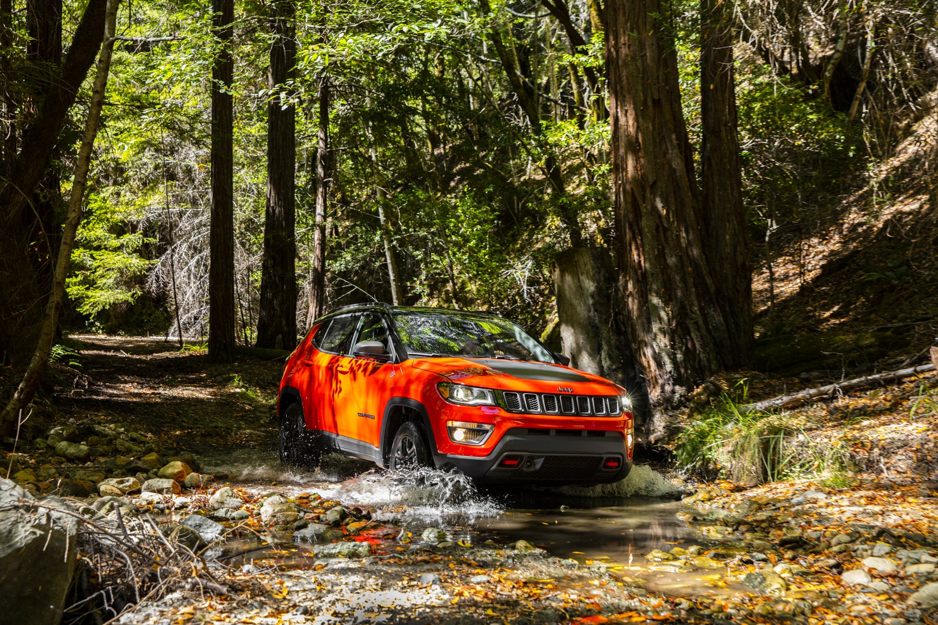 HD desktop wallpaper featuring a bright orange Jeep Trailhawk SUV driving through a forest stream, showcasing rugged off-road capability.