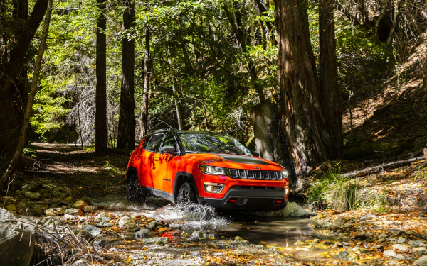 HD desktop wallpaper featuring a bright orange Jeep Trailhawk SUV driving through a forest stream, showcasing rugged off-road capability.