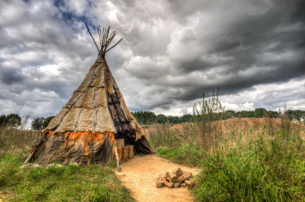 HDR image of a Native American man-made tipi under dramatic cloud-filled skies, set in a natural grassy landscape, captured as an HD desktop wallpaper.