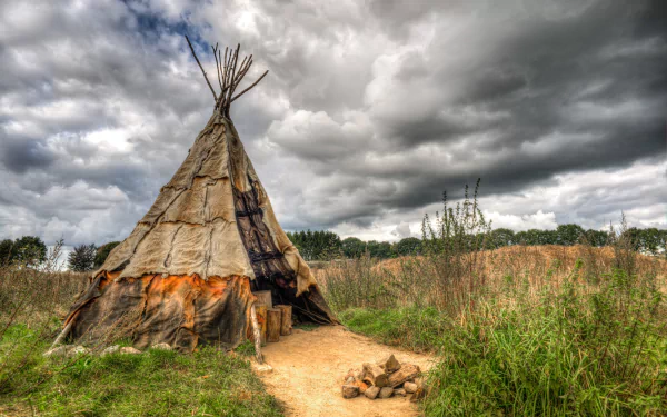 HDR image of a Native American man-made tipi under dramatic cloud-filled skies, set in a natural grassy landscape, captured as an HD desktop wallpaper.
