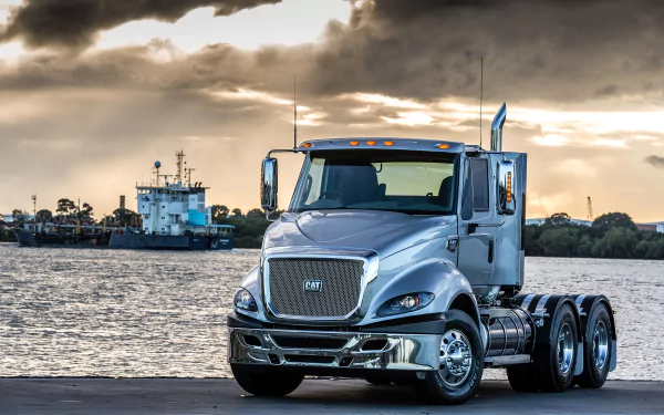 HD desktop wallpaper featuring a silver Caterpillar Inc. truck parked near a waterfront with boats and a dramatic cloudy sky in the background.