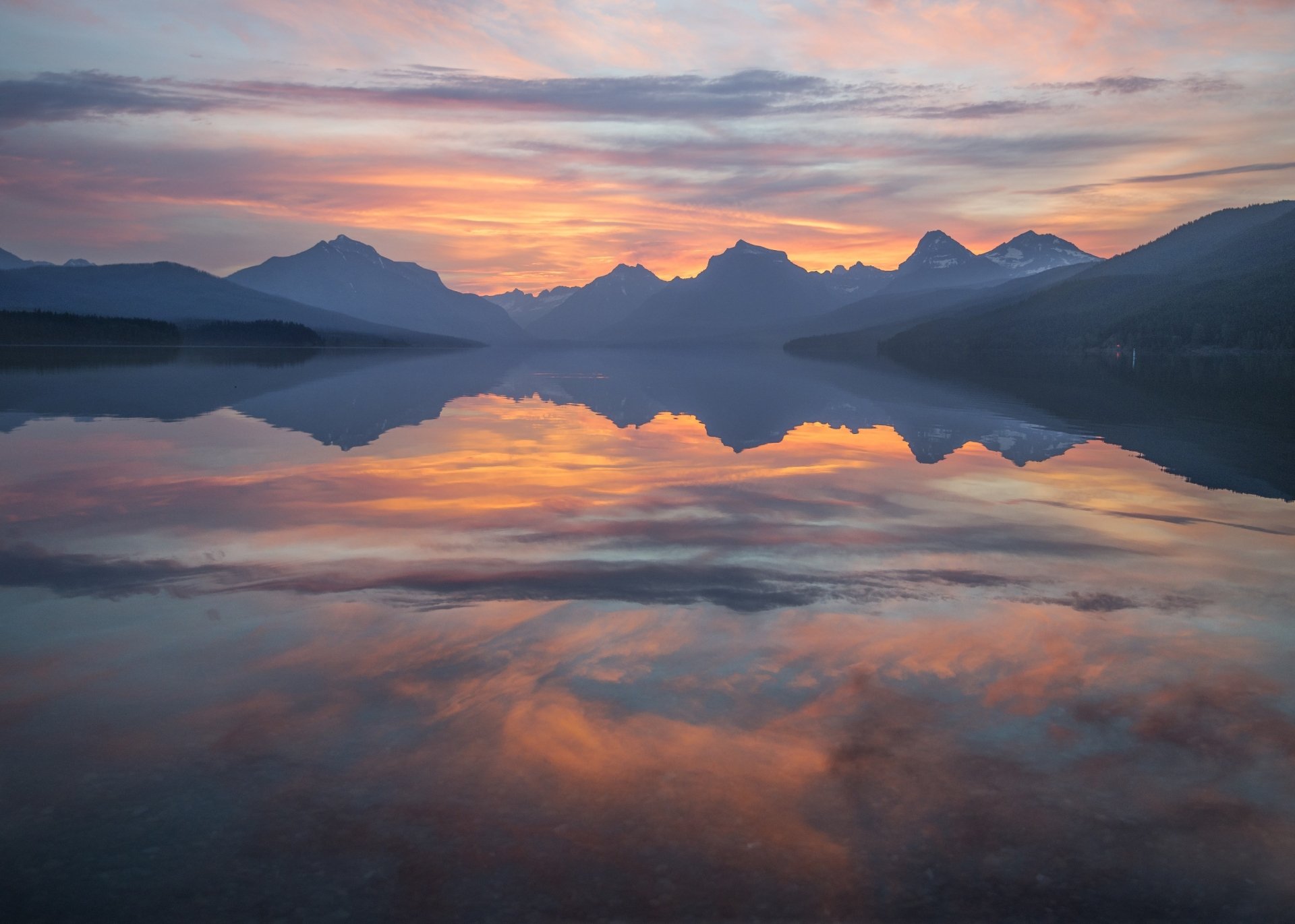 Sunrise over a mountain lake in Glacier National Park, Montana, USA, with clouds reflecting on the calm water at dawn.