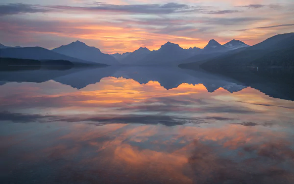 Sunrise over a mountain lake in Glacier National Park, Montana, USA, with clouds reflecting on the calm water at dawn.