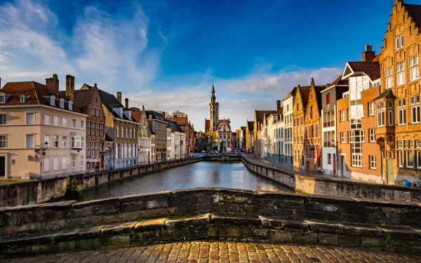Cityscape of Bruges, Belgium, featuring historic houses and buildings lining a canal with a stone bridge in the foreground under a vibrant blue sky.