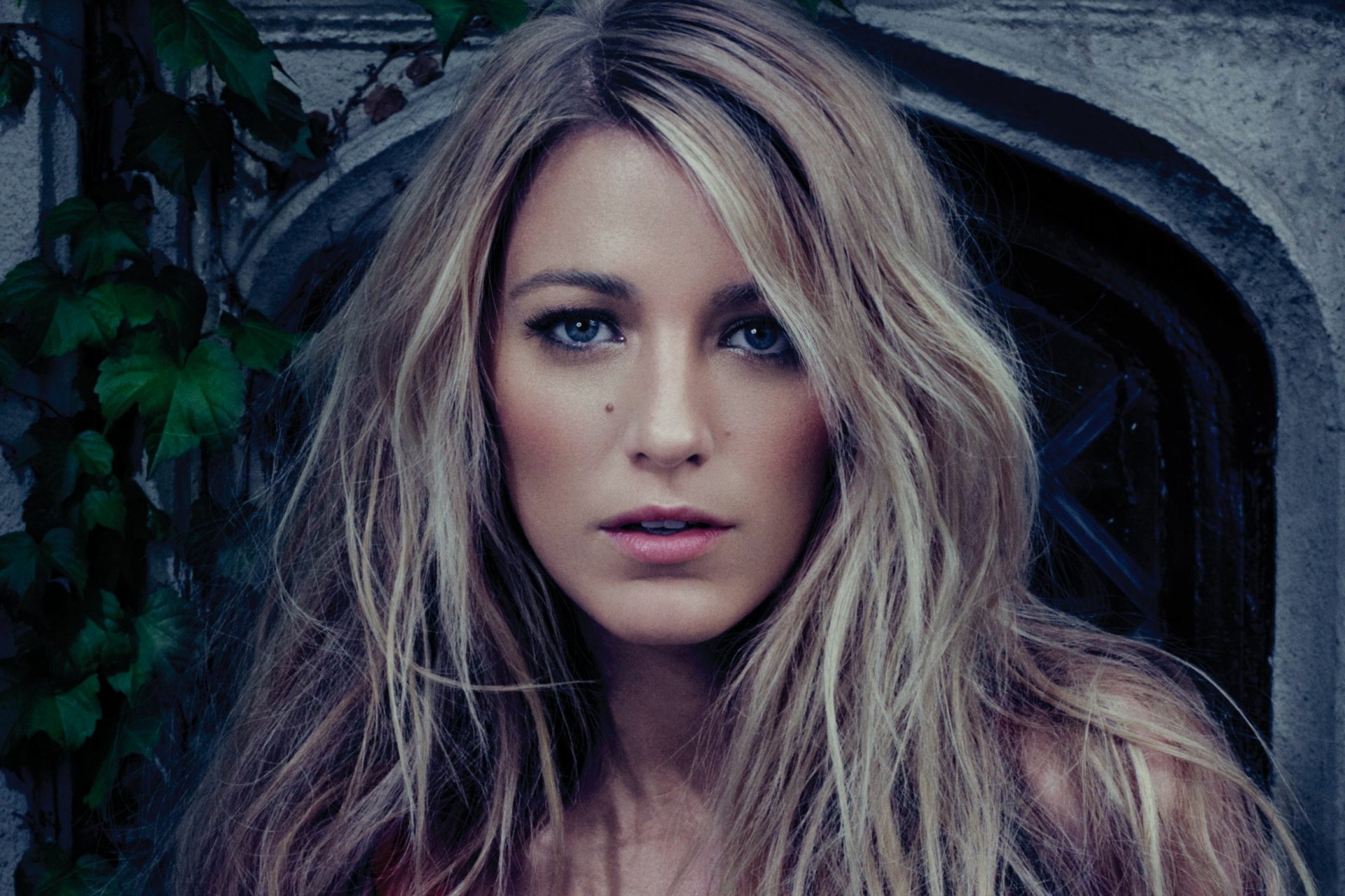 HD desktop wallpaper of blonde actress and celebrity Blake Lively with intense gaze, framed by natural foliage and a dark stone archway.
