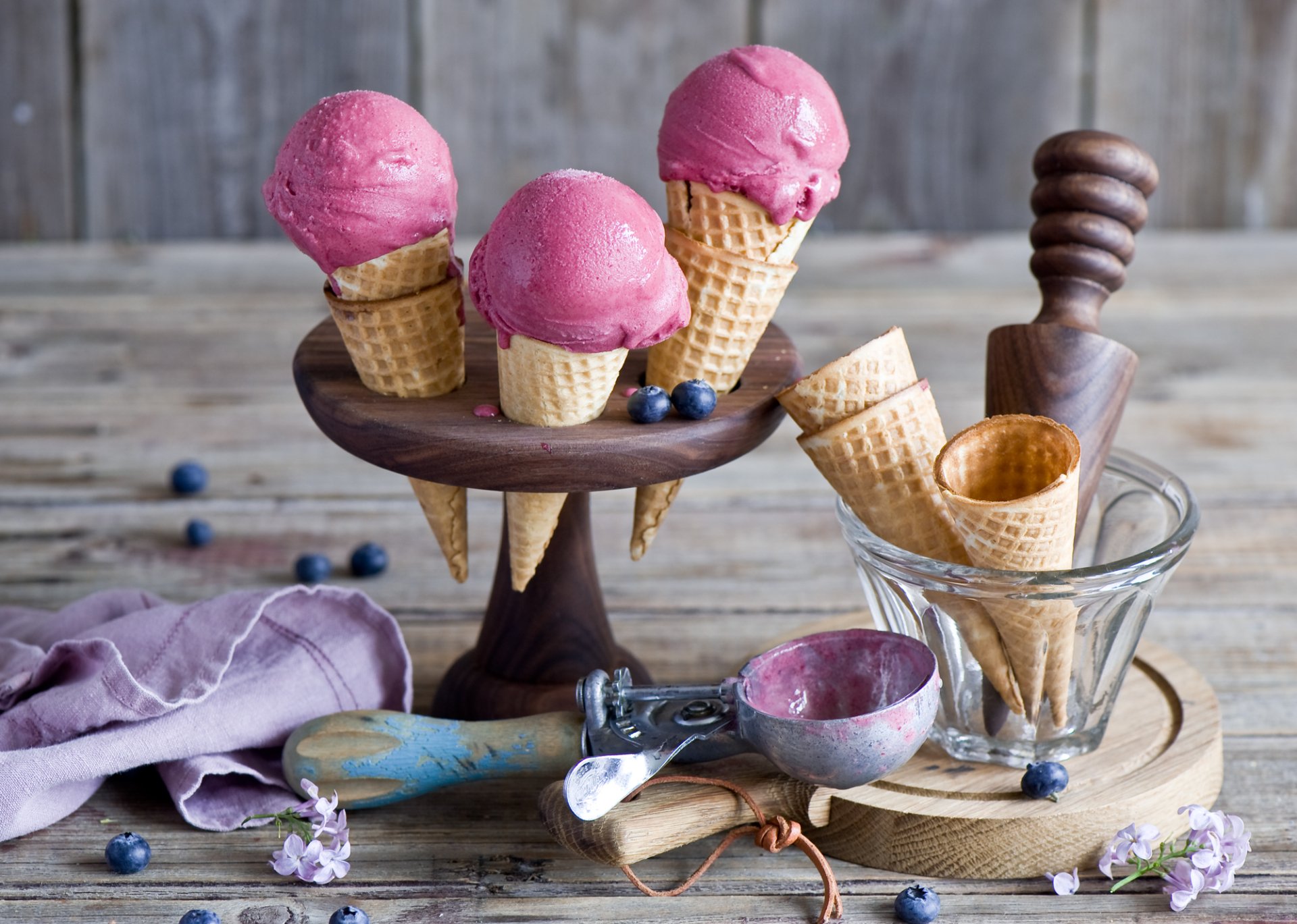 HD still life desktop wallpaper featuring three pink ice cream scoops in waffle cones on a wooden stand, with extra cones, an ice cream scoop, and blueberries on a rustic table.