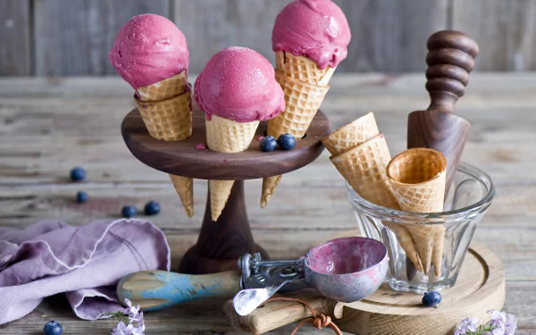HD still life desktop wallpaper featuring three pink ice cream scoops in waffle cones on a wooden stand, with extra cones, an ice cream scoop, and blueberries on a rustic table.