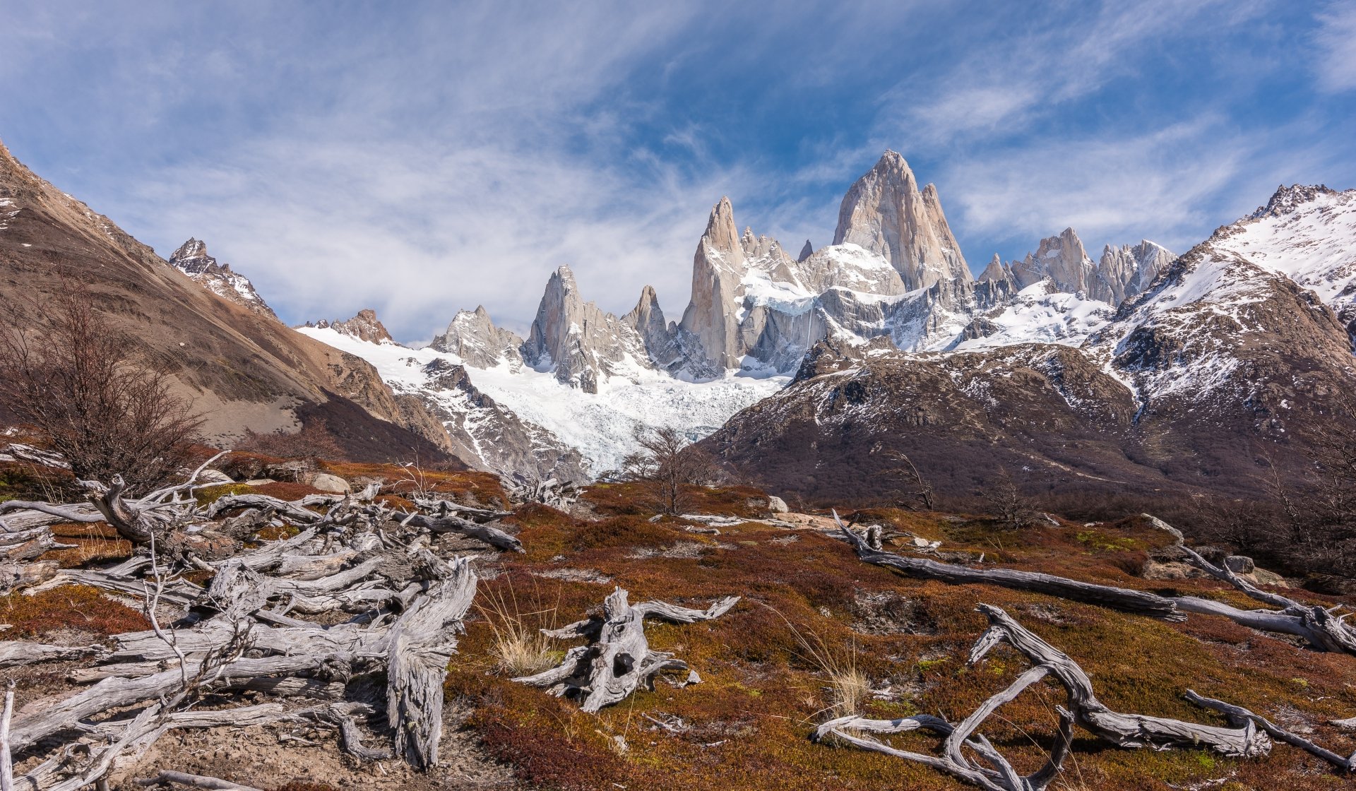 4K Ultra HD landscape of a rugged mountain range under a partly cloudy sky, with dry fallen trees in the foreground and snow-capped peaks in the background.