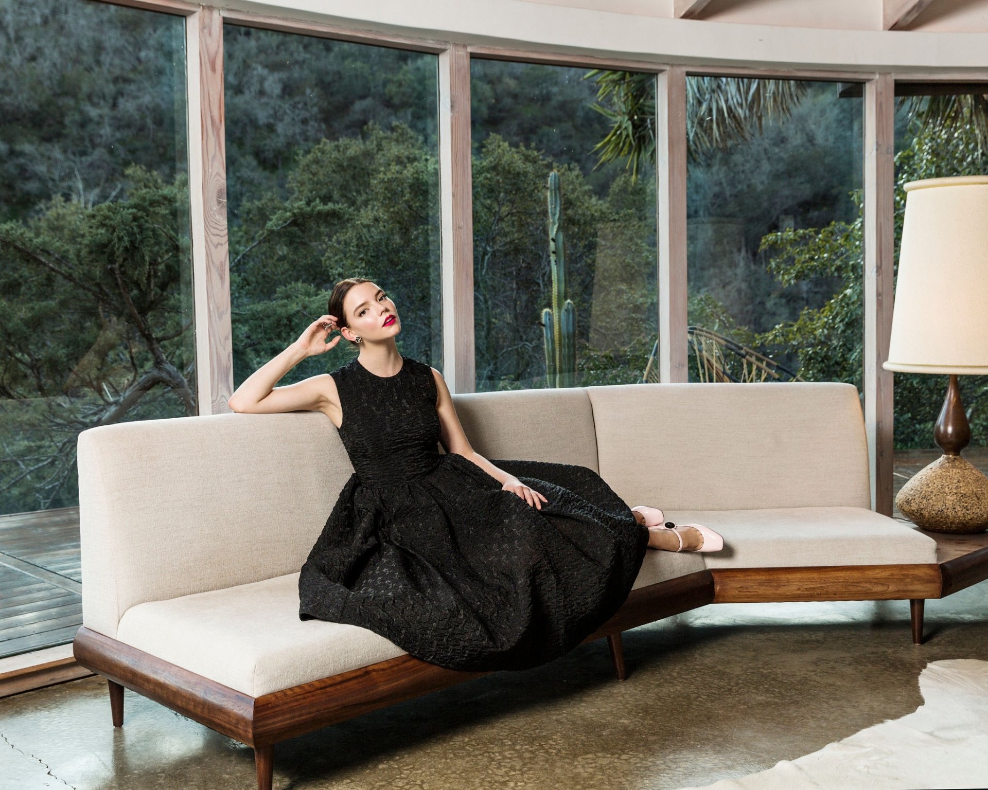 Brunette American actress and model Anya Taylor-Joy in a black dress with red lipstick lounging on a sofa by a large window in an HD desktop wallpaper background.