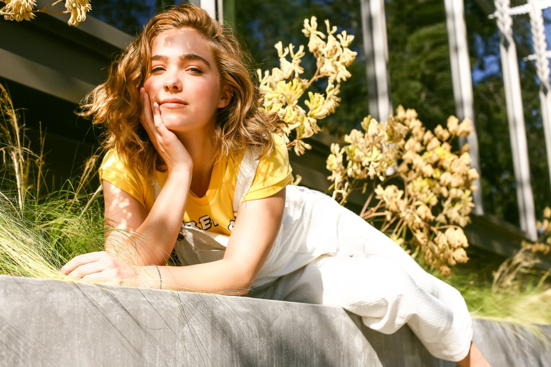 HD desktop wallpaper featuring American brunette actress Haley Lu Richardson reclining outdoors in natural sunlight with greenery and flowers in the background.