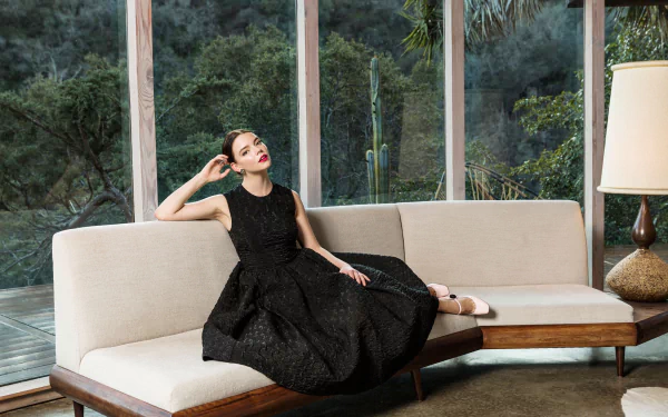 Brunette American actress and model Anya Taylor-Joy in a black dress with red lipstick lounging on a sofa by a large window in an HD desktop wallpaper background.