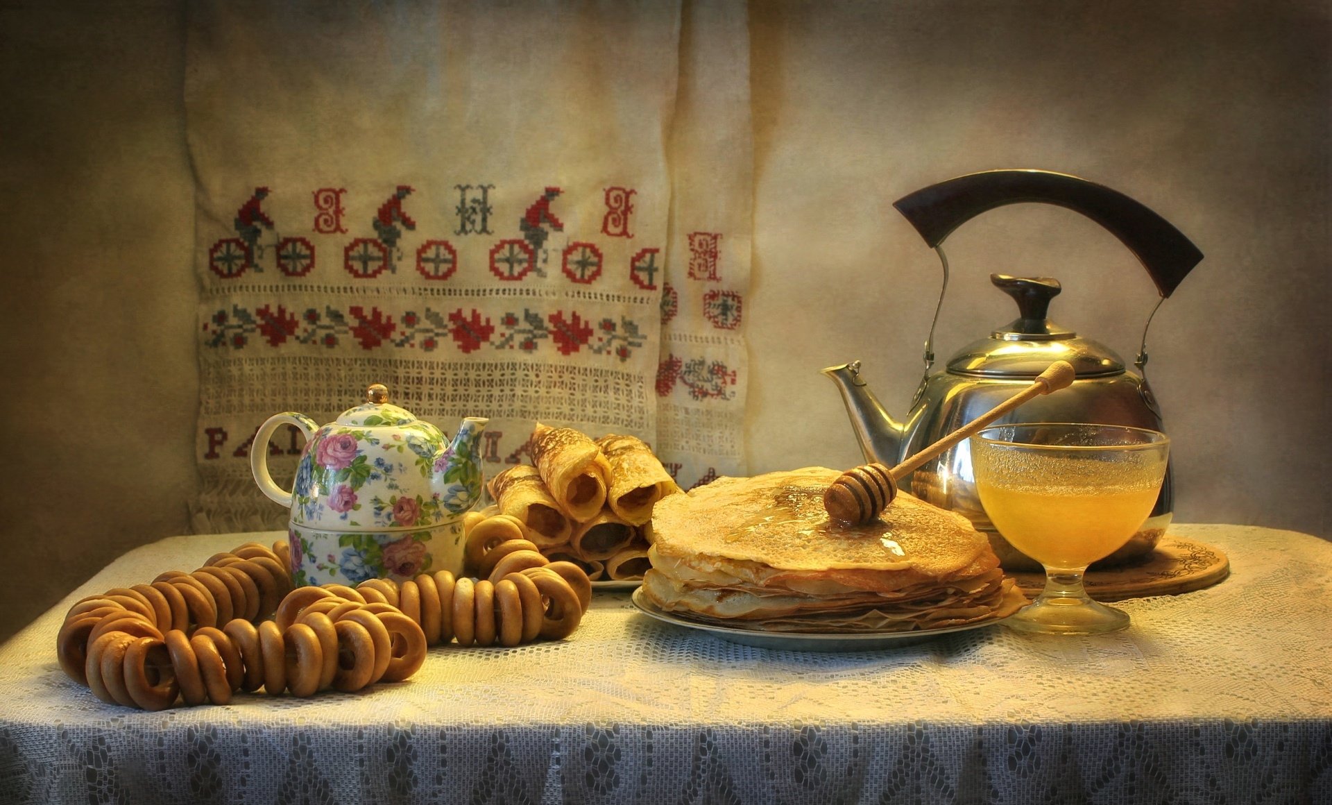 HD still life desktop wallpaper featuring crêpes with honey, a floral teapot, and stacked bagels on a textured table, illuminated by warm, soft lighting.